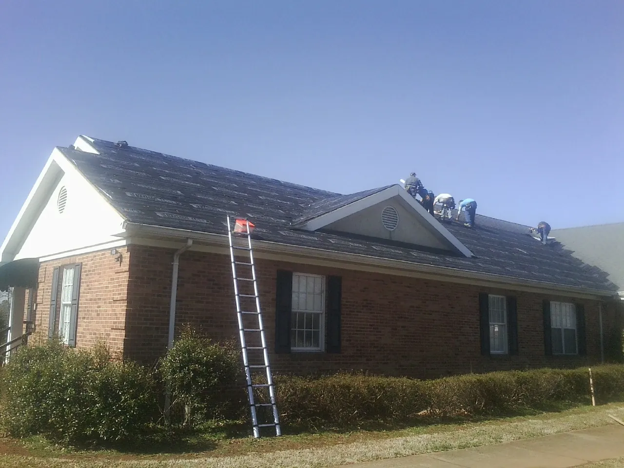 Crew tearing shingles off a brick ranch roof with a ladder set — Emergency Roof Repair work by our crew in White Hall