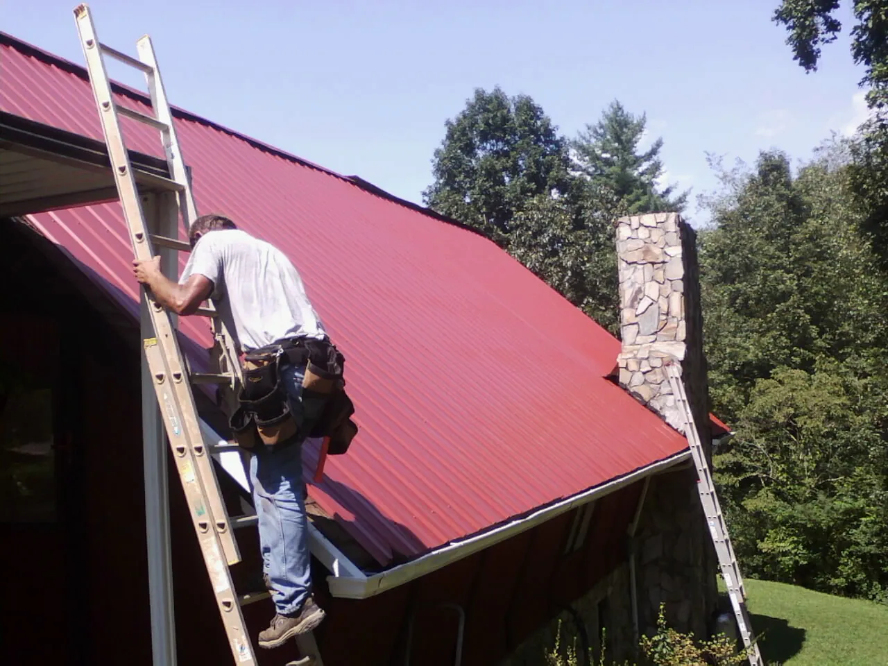 Roofer on a ladder beside a red metal roof and stone chimney — Soffit & Fascia Repair by Roofing Brothers in White Hall